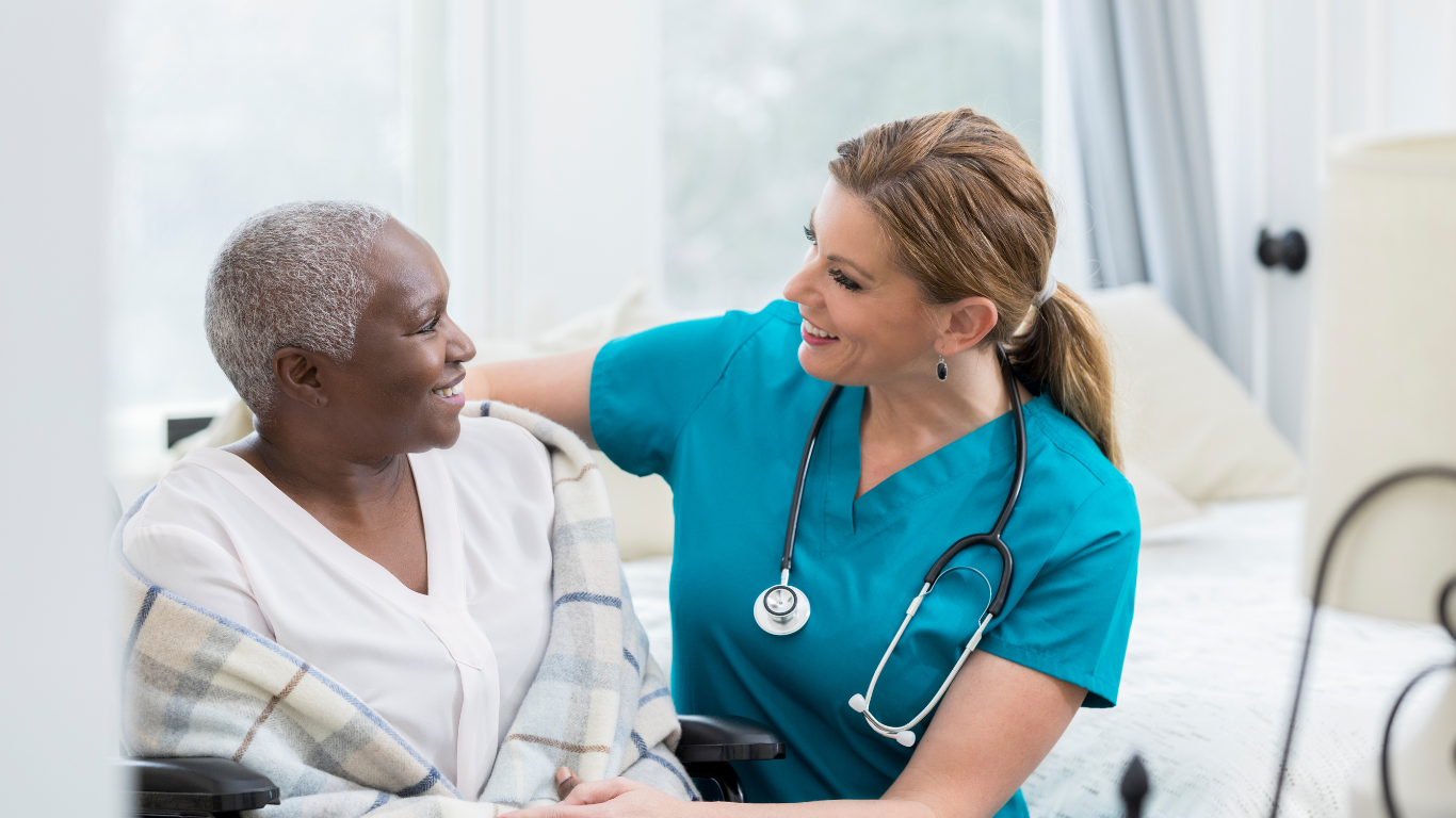 A health care worker sits next to a senior woman in a wheelchair and they smile at each other